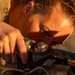 A person is closely inspecting or working on a mechanical component, leaning in with intense focus. The image is bathed in warm golden-hour light, creating dramatic shadows across the subject's face. The individual appears to be a technician or mechanic performing detailed hands-on work.