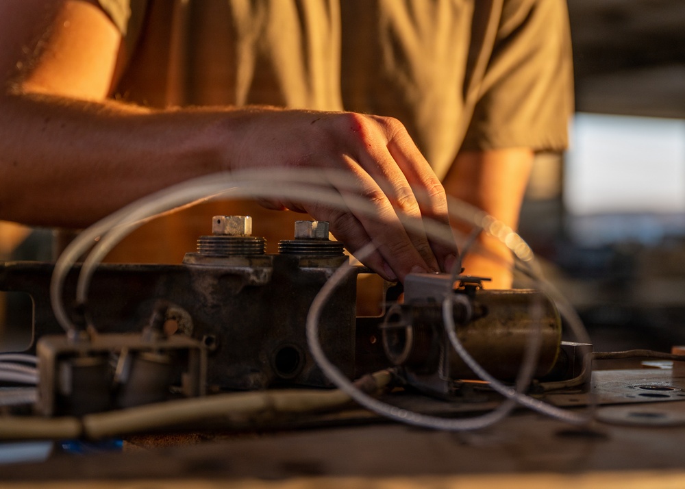 A person wearing an olive/tan t-shirt is working on a heavy mechanical component, likely an engine block or hydraulic manifold, placed on a flat metal surface. The component is heavily worn and dirty, with multiple threaded ports, fittings, and flexible hoses/lines attached. The scene is lit with warm golden light, suggesting either an outdoor setting during golden hour or a workshop with warm artificial lighting.