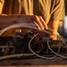 A person wearing an olive/tan t-shirt is working on a heavy mechanical component, likely an engine block or hydraulic manifold, placed on a flat metal surface. The component is heavily worn and dirty, with multiple threaded ports, fittings, and flexible hoses/lines attached. The scene is lit with warm golden light, suggesting either an outdoor setting during golden hour or a workshop with warm artificial lighting.