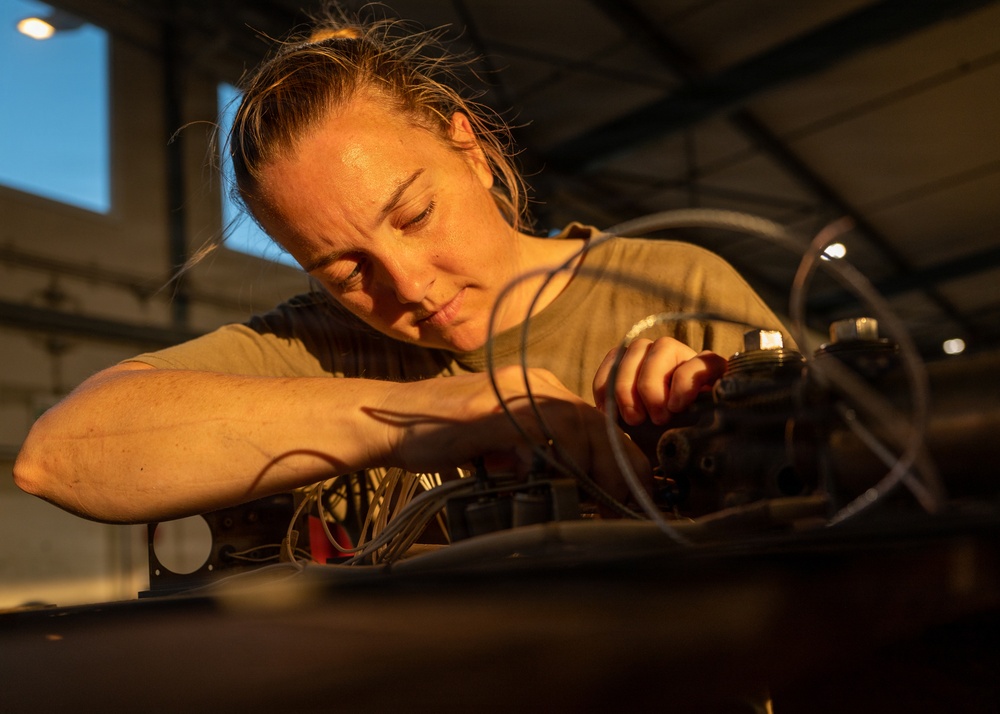 A woman is intently working on a complex assembly of wires, cables, and mechanical components in what appears to be a hangar or industrial workshop setting. She is leaning closely over the equipment, using her hands to manipulate or connect components. The image is dramatically lit with warm golden light highlighting her focused expression and work.