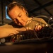 A woman is intently working on a complex assembly of wires, cables, and mechanical components in what appears to be a hangar or industrial workshop setting. She is leaning closely over the equipment, using her hands to manipulate or connect components. The image is dramatically lit with warm golden light highlighting her focused expression and work.