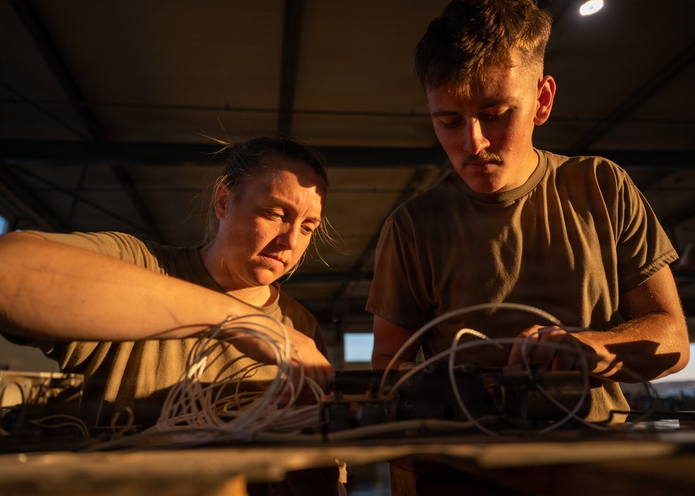 Two individuals wearing olive/military-green t-shirts are working together on a piece of equipment with multiple wires and cables on a workbench. They appear to be in an industrial or hangar-type structure with a metal roof, illuminated by warm golden light. Both are focused intently on the task, suggesting a technical or maintenance activity.