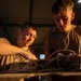 Two individuals wearing olive/military-green t-shirts are working together on a piece of equipment with multiple wires and cables on a workbench. They appear to be in an industrial or hangar-type structure with a metal roof, illuminated by warm golden light. Both are focused intently on the task, suggesting a technical or maintenance activity.