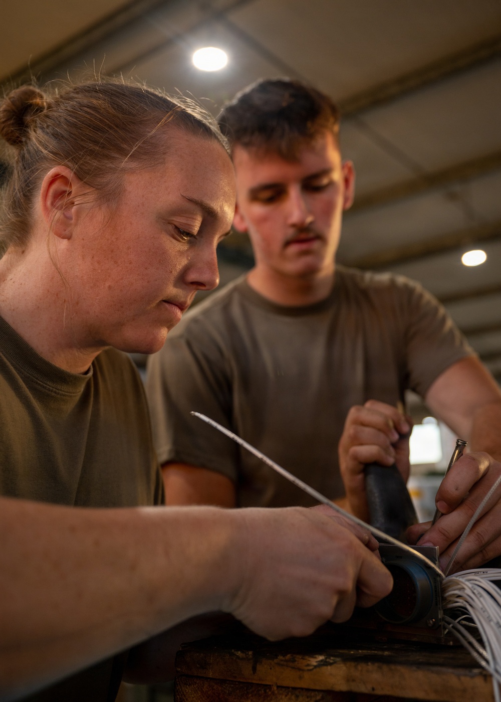 Two individuals wearing olive/military green t-shirts are working together on what appears to be an electrical or wiring task. The person in the foreground, a woman with her hair in a bun, is using a tool on a bundle of wires, while the man in the background observes and assists. The setting appears to be an indoor workshop or hangar with recessed ceiling lights.