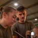 Two individuals wearing olive/military green t-shirts are working together on what appears to be an electrical or wiring task. The person in the foreground, a woman with her hair in a bun, is using a tool on a bundle of wires, while the man in the background observes and assists. The setting appears to be an indoor workshop or hangar with recessed ceiling lights.