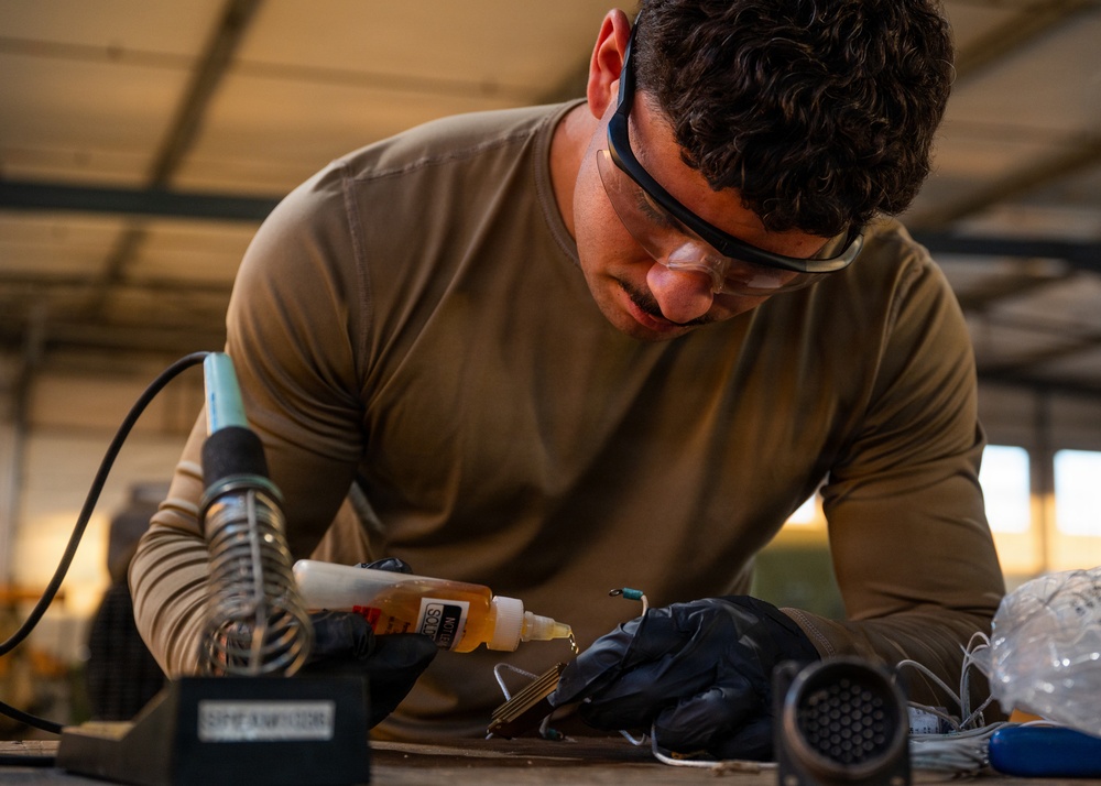A young man wearing safety glasses and black nitrile gloves is performing soldering or electrical work at a workbench in what appears to be a workshop or hangar. He is wearing an olive/tan long-sleeve shirt and is focused on working with small components, likely wiring or connectors. Various tools and supplies are visible on the workbench around him.
