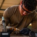 A young man wearing safety glasses and black nitrile gloves is performing soldering or electrical work at a workbench in what appears to be a workshop or hangar. He is wearing an olive/tan long-sleeve shirt and is focused on working with small components, likely wiring or connectors. Various tools and supplies are visible on the workbench around him.