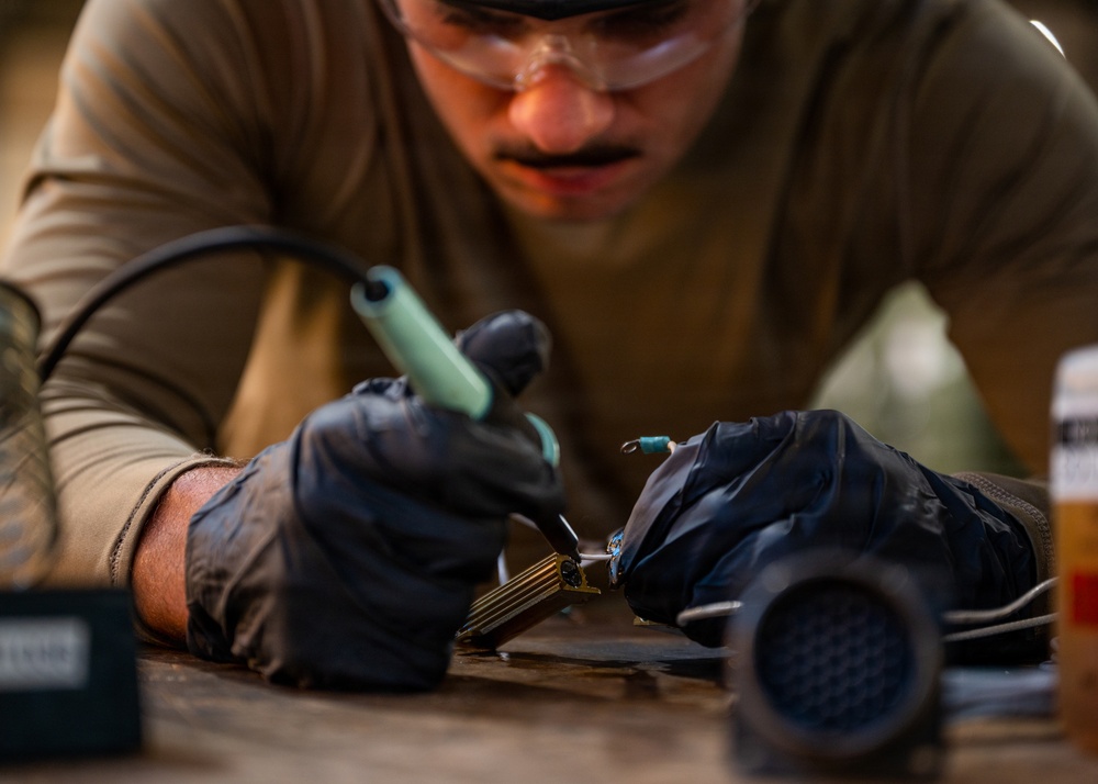 A person wearing safety goggles, black nitrile gloves, and an olive/tan long-sleeve shirt is performing close-up soldering or fine electronic work on a small component at a workbench. The individual is leaning in closely, indicating precision work requiring careful attention. Several tools and small containers are visible on the work surface nearby.