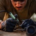 A person wearing safety goggles, black nitrile gloves, and an olive/tan long-sleeve shirt is performing close-up soldering or fine electronic work on a small component at a workbench. The individual is leaning in closely, indicating precision work requiring careful attention. Several tools and small containers are visible on the work surface nearby.