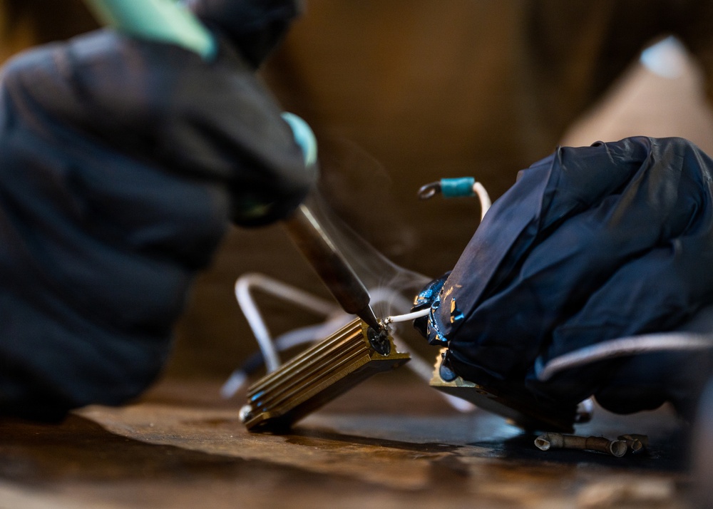 A close-up photograph shows a person wearing black nitrile gloves performing soldering work on an electronic component. Visible smoke rises from the soldering point, indicating active heat application to what appears to be a heat sink or electronic assembly with wires. The shallow depth of field keeps the focal point sharp while blurring the background.