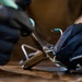 A close-up photograph shows a person wearing black nitrile gloves performing soldering work on an electronic component. Visible smoke rises from the soldering point, indicating active heat application to what appears to be a heat sink or electronic assembly with wires. The shallow depth of field keeps the focal point sharp while blurring the background.