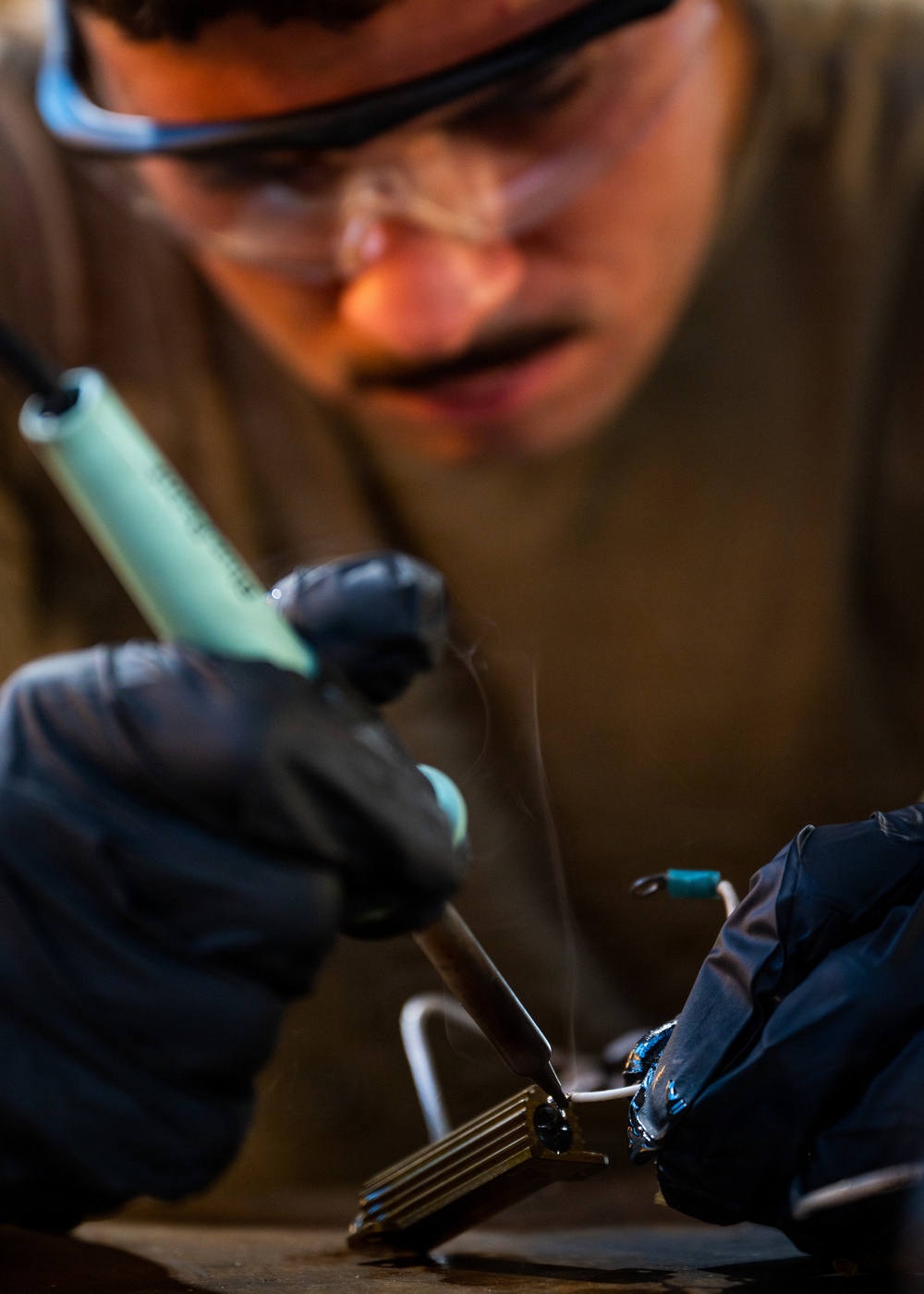 A person wearing safety goggles and black nitrile gloves is performing soldering work on an electronic component. Smoke/flux fumes are visibly rising from the soldering point, indicating active heat application. The image is a close-up shot with shallow depth of field, keeping the soldering action in sharp focus while the worker's face is slightly blurred in the background.