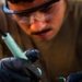 A person wearing safety goggles and black nitrile gloves is performing soldering work on an electronic component. Smoke/flux fumes are visibly rising from the soldering point, indicating active heat application. The image is a close-up shot with shallow depth of field, keeping the soldering action in sharp focus while the worker's face is slightly blurred in the background.