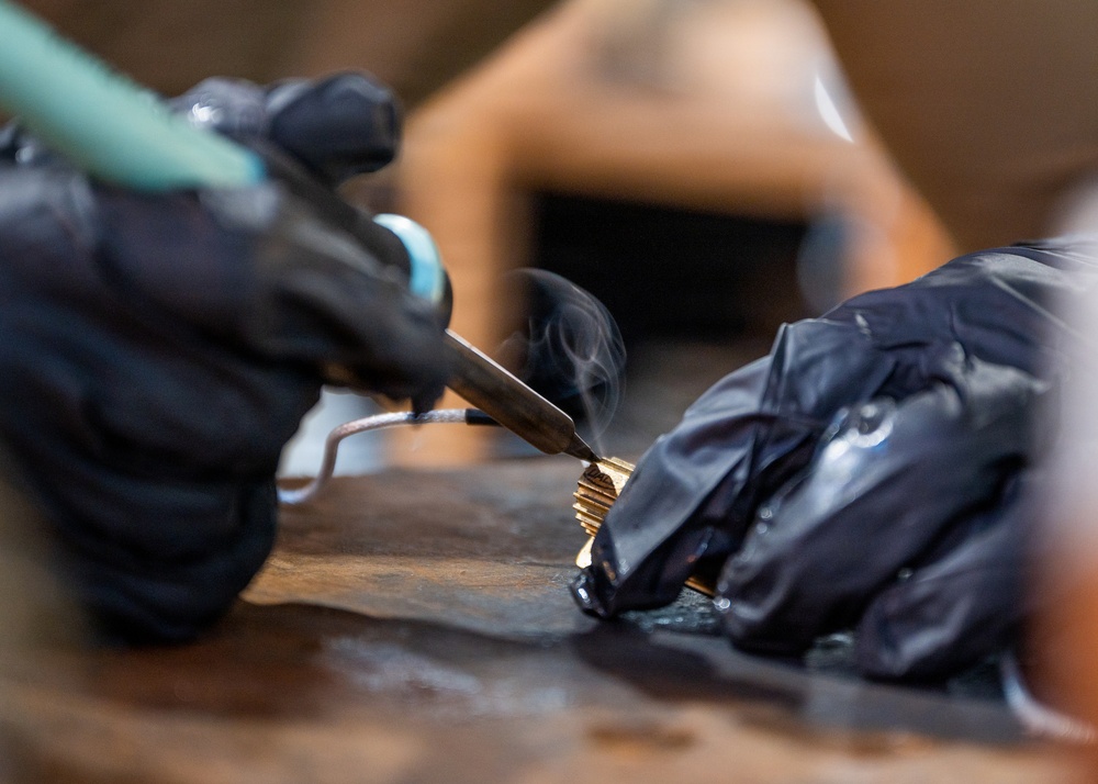 A person wearing black nitrile gloves is using a soldering iron or similar heat tool on a small gold/brass-colored metal component placed on a worn wooden surface. Visible smoke rises from the point of contact, indicating active heat application. The close-up macro shot captures fine detail of the hands and tool in a workshop or craft setting.
