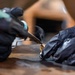 A person wearing black nitrile gloves is using a soldering iron or similar heat tool on a small gold/brass-colored metal component placed on a worn wooden surface. Visible smoke rises from the point of contact, indicating active heat application. The close-up macro shot captures fine detail of the hands and tool in a workshop or craft setting.
