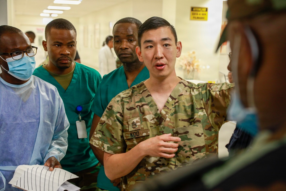 A U.S. Army soldier in camouflage uniform with the name tape 'KOO' and sergeant rank insignia appears to be briefing or instructing a group in what looks like a medical facility hallway. The group includes individuals wearing surgical scrubs and medical gowns, suggesting a military medical training or advisory mission. The setting appears to be a hospital or clinic, likely in an overseas location based on contextual clues.