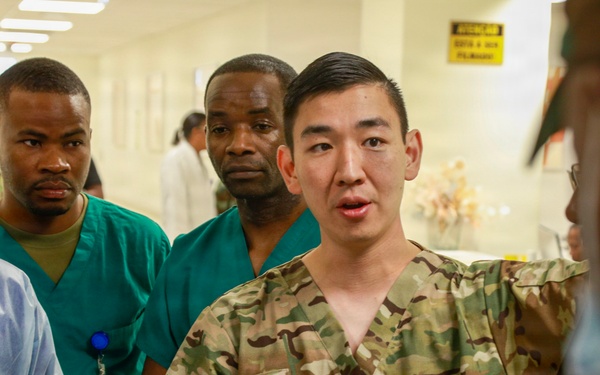 A U.S. Army soldier in camouflage uniform with the name tape 'KOO' and sergeant rank insignia appears to be briefing or instructing a group in what looks like a medical facility hallway. The group includes individuals wearing surgical scrubs and medical gowns, suggesting a military medical training or advisory mission. The setting appears to be a hospital or clinic, likely in an overseas location based on contextual clues.