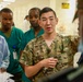 A U.S. Army soldier in camouflage uniform with the name tape 'KOO' and sergeant rank insignia appears to be briefing or instructing a group in what looks like a medical facility hallway. The group includes individuals wearing surgical scrubs and medical gowns, suggesting a military medical training or advisory mission. The setting appears to be a hospital or clinic, likely in an overseas location based on contextual clues.