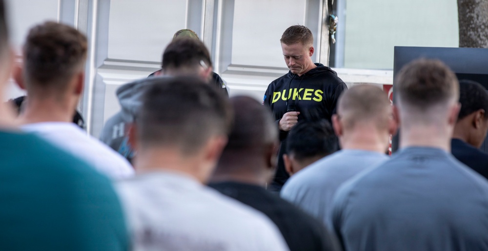 A man wearing a black hoodie with 'DUKES' printed in neon yellow lettering stands facing a group of people with their heads bowed. The setting appears to be outdoors near a building, and the atmosphere suggests a moment of reflection, prayer, or a group address. The central figure appears to be leading or speaking to the assembled group.