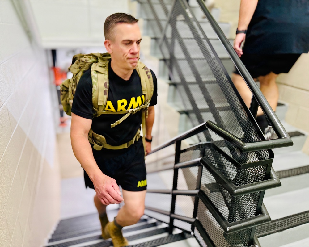 A young male soldier wearing a black U.S. Army PT (physical training) shirt and carrying a military rucksack is climbing an interior staircase. He appears to be engaged in a ruck march or stair-climbing fitness exercise, showing visible physical exertion. A second person in civilian clothing is partially visible ahead of him on the stairs.