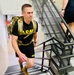 A young male soldier wearing a black U.S. Army PT (physical training) shirt and carrying a military rucksack is climbing an interior staircase. He appears to be engaged in a ruck march or stair-climbing fitness exercise, showing visible physical exertion. A second person in civilian clothing is partially visible ahead of him on the stairs.