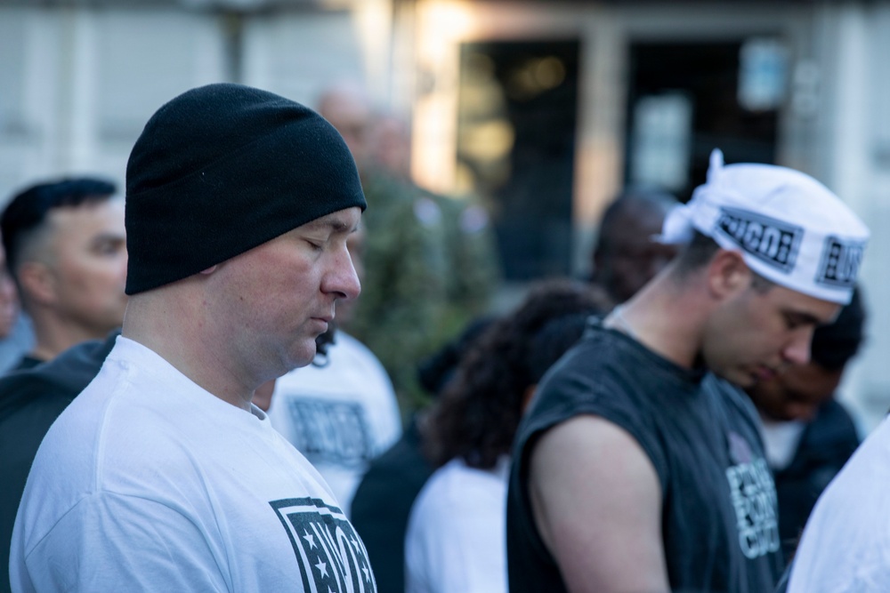 A group of people are gathered outdoors, with heads bowed in what appears to be a moment of reflection, prayer, or a group ceremony. Several individuals are wearing matching black-and-white branded apparel, suggesting an organized group event or fitness community gathering. The foreground features two prominent figures — one in a white t-shirt with a black beanie, and another in a black sleeveless shirt with a white branded headband.