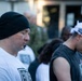 A group of people are gathered outdoors, with heads bowed in what appears to be a moment of reflection, prayer, or a group ceremony. Several individuals are wearing matching black-and-white branded apparel, suggesting an organized group event or fitness community gathering. The foreground features two prominent figures — one in a white t-shirt with a black beanie, and another in a black sleeveless shirt with a white branded headband.