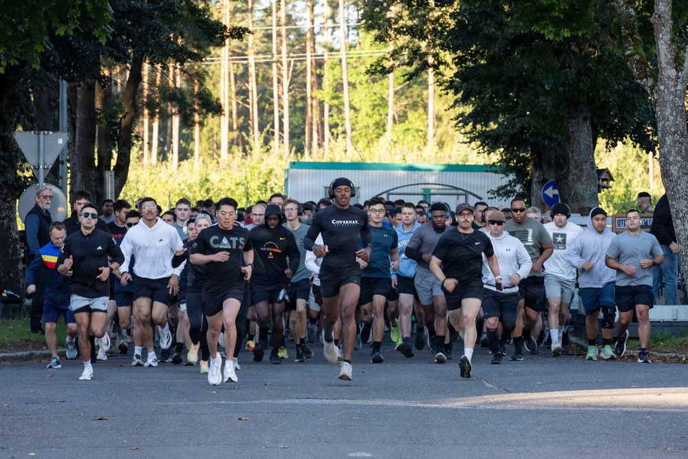 A large group of people are participating in an organized run or road race on a paved road lined with trees. The participants are dressed in casual athletic wear including t-shirts and shorts, suggesting a community or military fitness run. The setting appears to be a military installation based on visible U.S. Army signage in the background.
