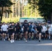 A large group of people are participating in an organized run or road race on a paved road lined with trees. The participants are dressed in casual athletic wear including t-shirts and shorts, suggesting a community or military fitness run. The setting appears to be a military installation based on visible U.S. Army signage in the background.