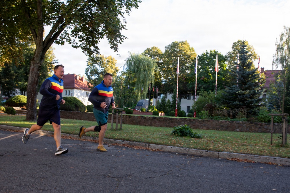Two middle-aged men are jogging together along a paved road in what appears to be a well-maintained institutional or military compound. Both are wearing matching blue athletic jackets with yellow and red horizontal stripes, suggesting they may be part of the same team or organization. The setting includes manicured grounds, flagpoles with flags, and residential-style buildings in the background.