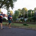 Two middle-aged men are jogging together along a paved road in what appears to be a well-maintained institutional or military compound. Both are wearing matching blue athletic jackets with yellow and red horizontal stripes, suggesting they may be part of the same team or organization. The setting includes manicured grounds, flagpoles with flags, and residential-style buildings in the background.