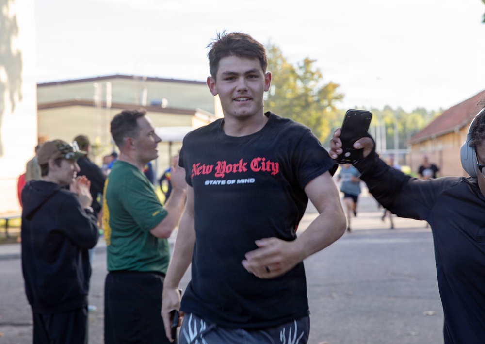 A young man wearing a black 'New York City State of Mind' t-shirt is running or jogging through what appears to be an outdoor event or race. Bystanders are cheering and someone is recording him with a smartphone. The setting appears to be a paved area near buildings, likely a military base or institutional campus.