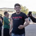 A young man wearing a black 'New York City State of Mind' t-shirt is running or jogging through what appears to be an outdoor event or race. Bystanders are cheering and someone is recording him with a smartphone. The setting appears to be a paved area near buildings, likely a military base or institutional campus.