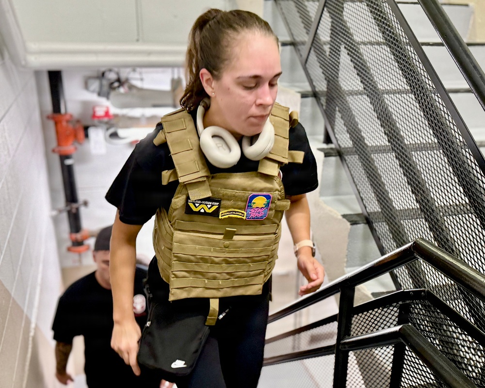 A young woman wearing a tan/coyote-colored tactical plate carrier vest over a black t-shirt is ascending a metal staircase in what appears to be an industrial or institutional building. She has white over-ear headphones around her neck and is carrying a small black Nike crossbody bag. A second person in a black cap is visible behind her on the stairs.