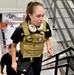 A young woman wearing a tan/coyote-colored tactical plate carrier vest over a black t-shirt is ascending a metal staircase in what appears to be an industrial or institutional building. She has white over-ear headphones around her neck and is carrying a small black Nike crossbody bag. A second person in a black cap is visible behind her on the stairs.