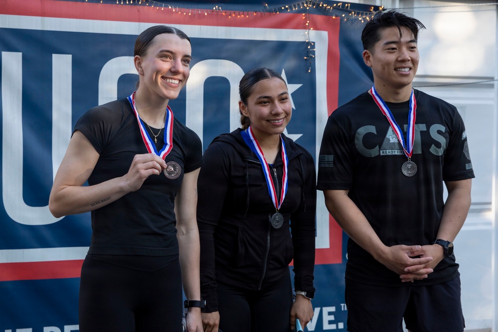 Three individuals wearing black athletic clothing stand together in front of a red, white, and blue banner, each displaying medals on red-white-and-blue ribbons around their necks. The leftmost person is holding their medal up for display while smiling broadly. The setting appears to be an awards ceremony or competition podium moment.