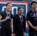 Three individuals wearing black athletic clothing stand together in front of a red, white, and blue banner, each displaying medals on red-white-and-blue ribbons around their necks. The leftmost person is holding their medal up for display while smiling broadly. The setting appears to be an awards ceremony or competition podium moment.