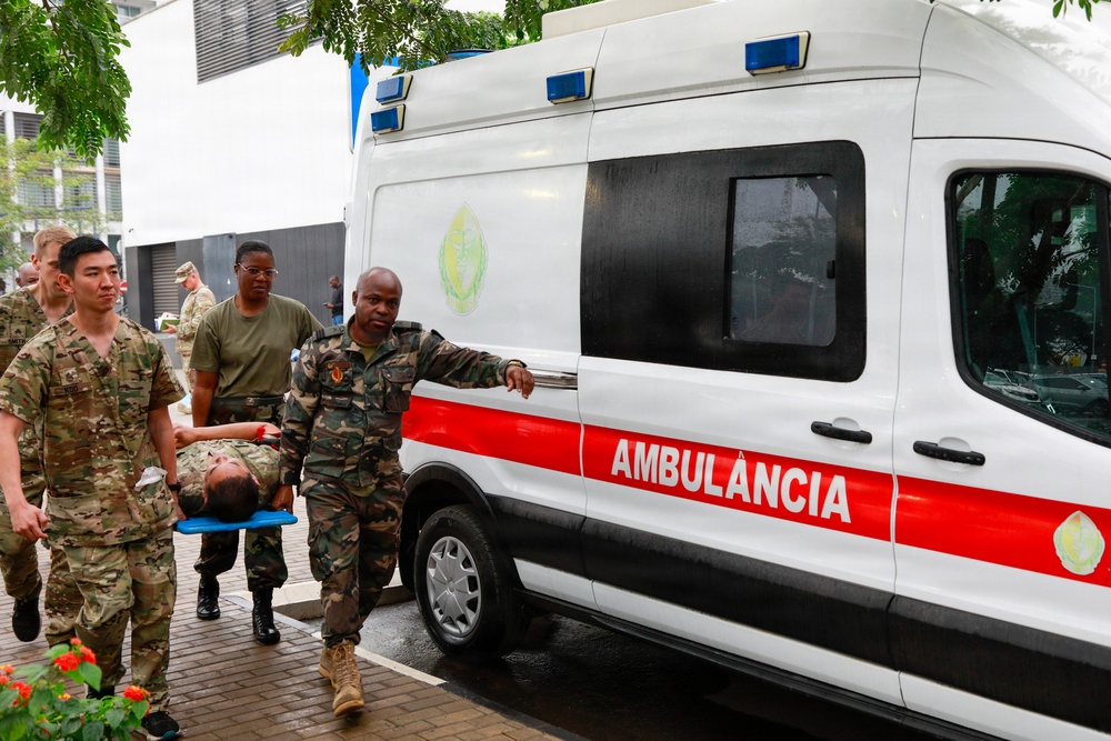 Military personnel in camouflage uniforms are carrying a person on a stretcher toward a white and red ambulance labeled 'AMBULÂNCIA'. The scene appears to be a medical evacuation or training exercise involving both U.S. and foreign military forces. The setting is an outdoor paved area near a building, under overcast/rainy conditions.
