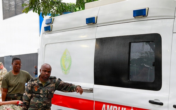 Military personnel in camouflage uniforms are carrying a person on a stretcher toward a white and red ambulance labeled 'AMBULÂNCIA'. The scene appears to be a medical evacuation or training exercise involving both U.S. and foreign military forces. The setting is an outdoor paved area near a building, under overcast/rainy conditions.