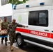 Military personnel in camouflage uniforms are carrying a person on a stretcher toward a white and red ambulance labeled 'AMBULÂNCIA'. The scene appears to be a medical evacuation or training exercise involving both U.S. and foreign military forces. The setting is an outdoor paved area near a building, under overcast/rainy conditions.