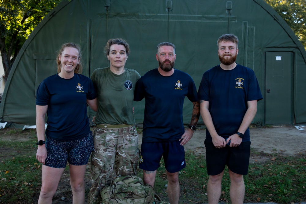 Four individuals pose together in front of a large olive-green military-style inflatable or modular tent structure. Three are wearing navy blue t-shirts with visible military unit insignia, while one wears an olive/green t-shirt with camouflage trousers. The setting appears to be a military base or field camp environment.