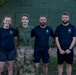 Four individuals pose together in front of a large olive-green military-style inflatable or modular tent structure. Three are wearing navy blue t-shirts with visible military unit insignia, while one wears an olive/green t-shirt with camouflage trousers. The setting appears to be a military base or field camp environment.
