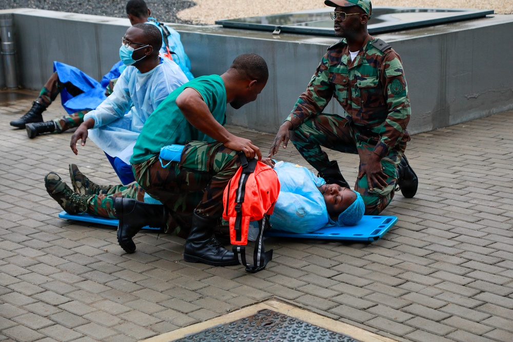 The image shows what appears to be a medical emergency drill or training exercise involving military personnel and medical staff. Two individuals in camouflage uniforms are assisting a person lying on a blue stretcher/backboard on a paved outdoor surface. Additional personnel in medical scrubs and protective gowns are visible in the background performing similar activities.