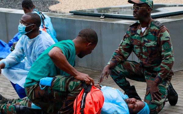 The image shows what appears to be a medical emergency drill or training exercise involving military personnel and medical staff. Two individuals in camouflage uniforms are assisting a person lying on a blue stretcher/backboard on a paved outdoor surface. Additional personnel in medical scrubs and protective gowns are visible in the background performing similar activities.