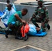 The image shows what appears to be a medical emergency drill or training exercise involving military personnel and medical staff. Two individuals in camouflage uniforms are assisting a person lying on a blue stretcher/backboard on a paved outdoor surface. Additional personnel in medical scrubs and protective gowns are visible in the background performing similar activities.