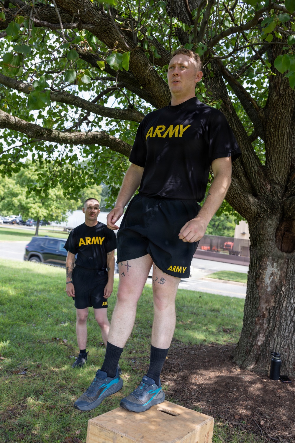 Two individuals wearing matching U.S. Army physical training (PT) uniforms — black shirts and shorts with gold 'ARMY' lettering — are outdoors on a grassy area beneath a large tree. The foreground subject is standing elevated on a wooden plyo box, while a second person stands on the ground behind them. The setting appears to be a military installation based on the uniforms and surrounding infrastructure.