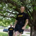 Two individuals wearing matching U.S. Army physical training (PT) uniforms — black shirts and shorts with gold 'ARMY' lettering — are outdoors on a grassy area beneath a large tree. The foreground subject is standing elevated on a wooden plyo box, while a second person stands on the ground behind them. The setting appears to be a military installation based on the uniforms and surrounding infrastructure.