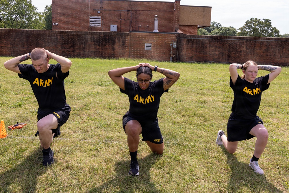 Three U.S. Army soldiers in matching black and gold PT (physical training) uniforms are performing lunges on a grass field. All three have their hands interlocked behind their heads, indicating a standardized exercise drill. The setting appears to be a military installation, with a brick building and wall visible in the background.