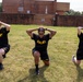 Three U.S. Army soldiers in matching black and gold PT (physical training) uniforms are performing lunges on a grass field. All three have their hands interlocked behind their heads, indicating a standardized exercise drill. The setting appears to be a military installation, with a brick building and wall visible in the background.