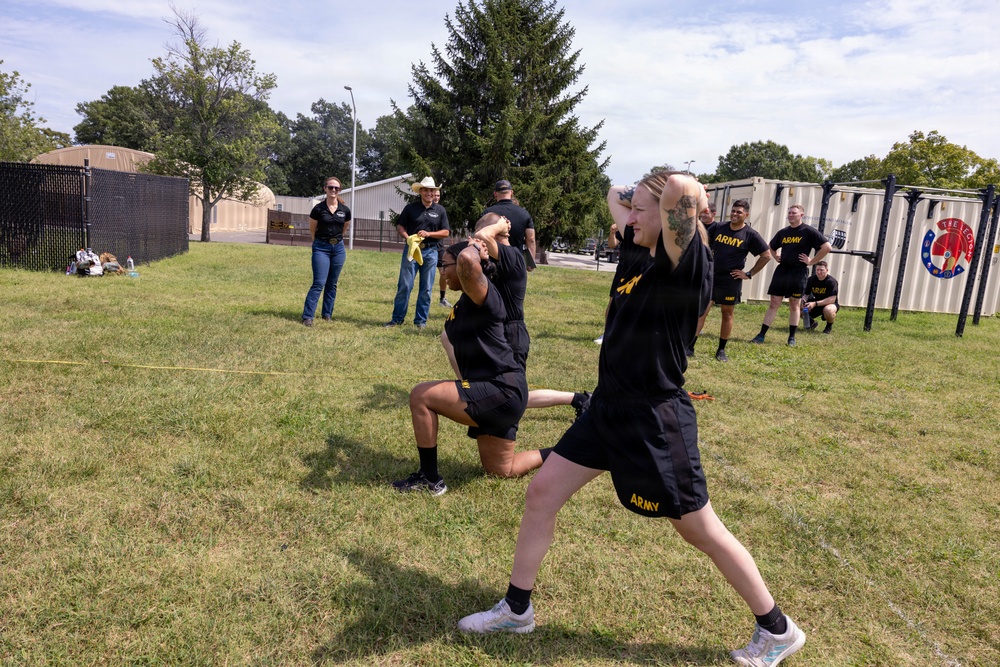 The image shows U.S. Army personnel conducting physical training exercises on a grass field. Two soldiers in the foreground wearing black Army PT (Physical Training) uniforms are performing lunges or stretching exercises with hands behind their heads. Several observers, including civilians and other soldiers, watch from the background near shipping containers and a chain-link fence.